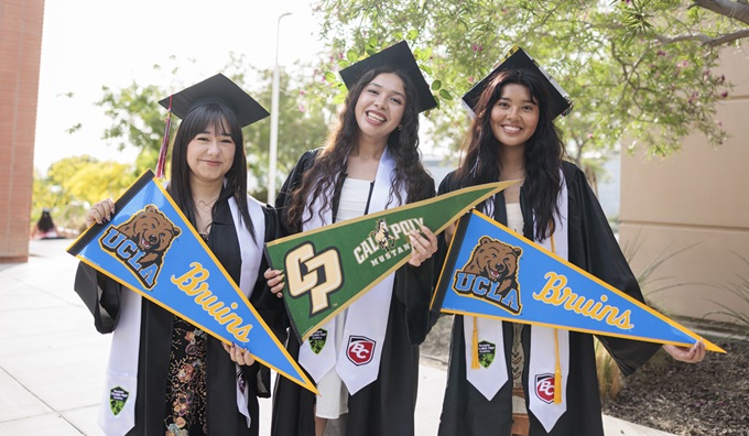 Three young people stand at a high school courtyard wearing graduation gowns and and showing university pennants