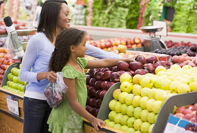 What looks like a mother and a daughter stand in front of a lemon display at the grocery store while choosing lemons at the produce aisle